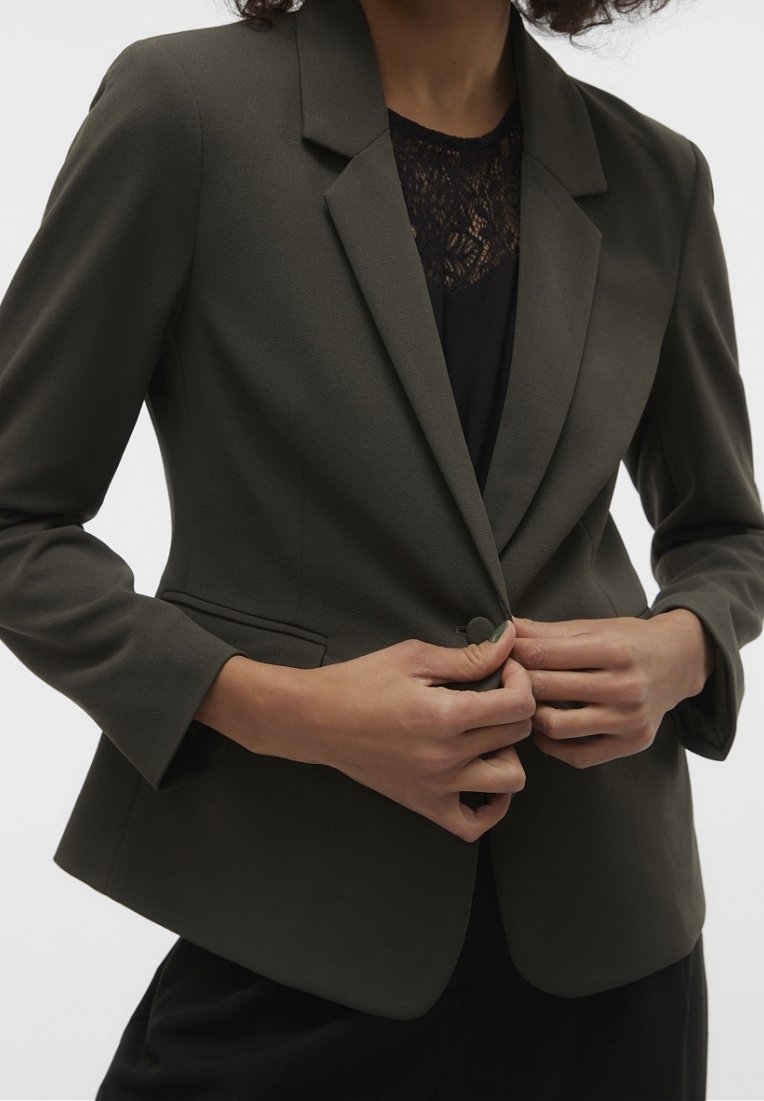 Des mains ferment un bouton sur un blazer vert olive foncé porté au-dessus d'un top en dentelle noire et d'un pantalon noir sur une personne aux cheveux bouclés.