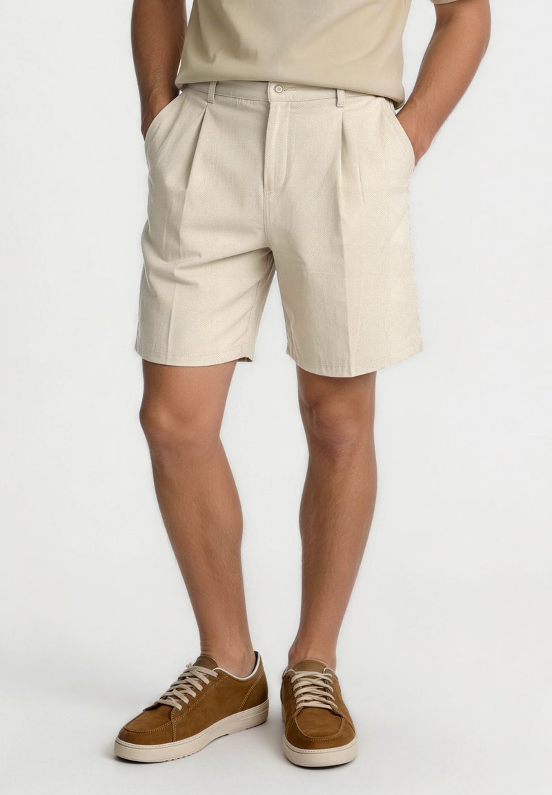 Short beige plissé porté avec une chemise assortie et des baskets marron à lacets, personne debout les mains dans les poches.