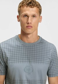 Ljusblå kortärmad t-shirt med rutmönster, rund halsringning och broderad logotyp på bröstet. Slät bomullstyg.