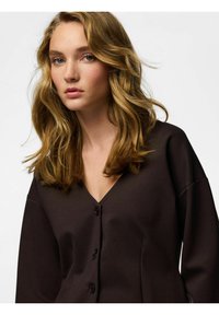 Tamno smeđa košulja s gumbima, V-izrezom i širokim rukavima, izrađena od glatkog tkanja; ima tri gumba sprijeda.