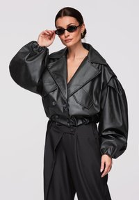 Vrouw draagt een oversized zwarte leren jas met opgeblazen mouwen en hooggetailleerde zwarte broek, terwijl ze smalle zwarte zonnebril rechtzet.