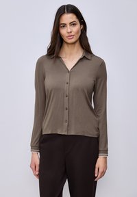 Femme aux longs cheveux foncés portant une chemise marron à manches longues boutonnée et un pantalon foncé, debout devant un fond clair uni.