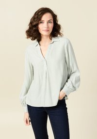 Hellgrün gestreifte Bluse mit Kragen, langen Ärmeln und Knopfleiste. Weiches Material mit lockerem Schnitt und abgerundetem Saum. Kombiniert mit dunklen Jeans.