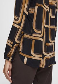 Camicia nera con motivi geometrici curvi in oro e marrone, texture setosa, maniche lunghe con bottone e orlo curvo. Pantaloni marroni visibili.