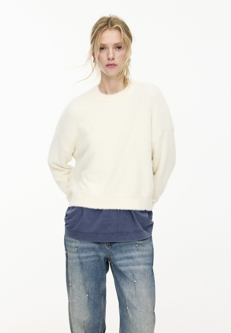 Pull en crème, moelleux, avec un col rond, associé à un bas superposé bleu marine et un jean bleu clair orné de clous.
