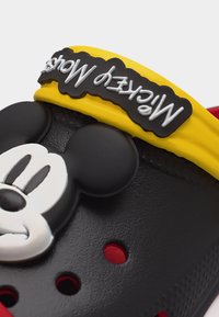 Μαύρο και κόκκινο παπούτσι με διακόσμηση το πρόσωπο του Μίκυ Μάους και κίτρινο λουράκι που φέρει το λογότυπο του Μίκυ Μάους σε λευκό κείμενο.