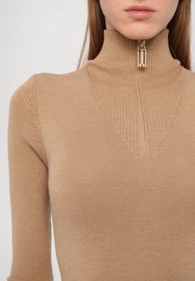 Col roulé en ribbed beige avec une fermeture à demi-zip. Présente un décolleté en forme de V et une texture lisse, mettant en valeur une silhouette ajustée.