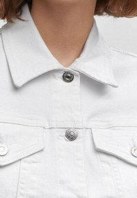 Giacca in denim bianco con colletto classico, bottoni a pressione argentati e tasche sul petto. Il tessuto appare robusto con una texture liscia.