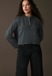 Grå beskåret langærmet top med rund halsudskæring og knaplukning, parret med sorte jeans med et subtilt zebra mønster.