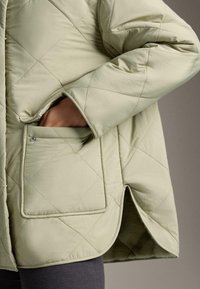 Svetlozelená prešívaná bunda so zapínaním na cvoky na vrecku, rukou vo vrecku a s zakriveným zakončením pod pazuchou cez tmavé nohavice.