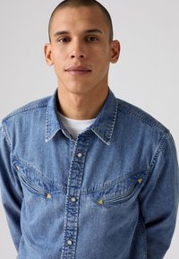 Denim overhemd in lichtblauw, met een puntige kraag, drukknopen en twee borstmouwen met driehoekige accenten. Zachte textuur.