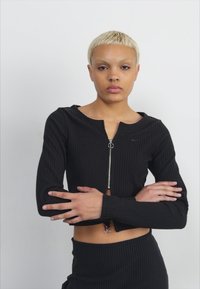Svart ribbet langermet crop topp med glidelås og rund hals. Har en liten Nike-logo på venstre bryst.