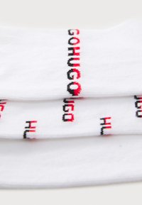 Weiße Baumwollsocken mit auffälligem, besticktem Text in Schwarz und Rot. Der Text beinhaltet "GO" und "HUGO" in einem sich wiederholenden Muster. Glatte Textur.