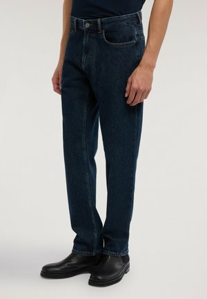 Man som bär mörkblå jeans med raka ben, svarta läderstövlar och en instoppad svart skjorta, stående mot en enfärgad bakgrund.