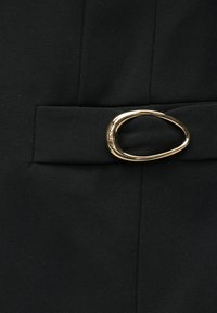 Μαύρο ύφασμα με λεία υφή που διαθέτει ορθογώνια ζώνη μέσης και λεπτομέρεια σε σχήμα οβάλ πόρπης σε χρυσό χρώμα.