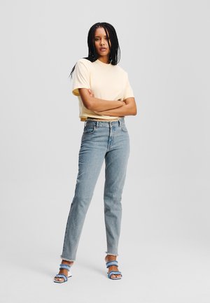 Camiseta amarilla recortada, jeans de tiro alto en azul claro y sandalias azules de múltiples correas. Texturas suaves y un ajuste casual.