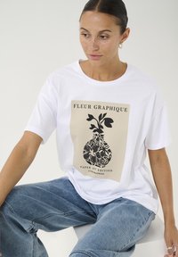 White short-sleeve t-shirt featuring a graphic print of a vase with leaves in black on a beige background, paired with light blue jeans.