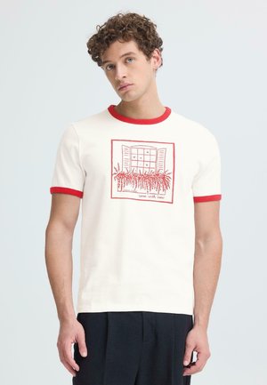 Młody mężczyzna z kręconymi włosami, ubrany w białą koszulkę z czerwonymi wykończeniami oraz nadrukiem czerwonego okna i rośliny, stojący na jednolitym jasnym tle.