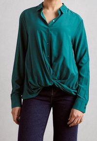 Blusa turchese con chiusura a bottone, maniche lunghe e dettaglio annodato in vita, realizzata in un tessuto morbido. Abbinata a jeans scuri.