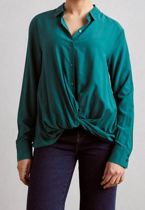 Türkise Bluse mit durchgehender Knopfleiste, langen Ärmeln und einem Knoten-Detail an der Taille, aus einem glatten Stoff gefertigt. Kombiniert mit dunklen Jeans.