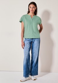 Top corto verde menta con collo henley e dettagli con bottoni, abbinato a jeans a gamba larga blu chiaro e sneakers bianche da ginnastica.