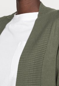 Olivengrøn strikket cardigan med ribbet tekstur, åbent frontdesign, lagdelt over en hvid t-shirt med lille brystlomme.