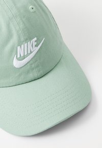 Világos zöld pamut sapka ívelt karimával, fehér hímzett Nike logóval és textúrázott anyag részletekkel.