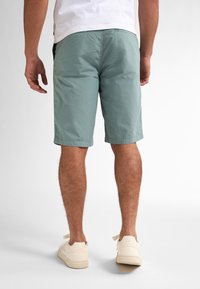 Shorts en coton vert clair avec un devant plat, une coupe droite et des poches latérales, associés à des baskets blanc cassé, vus de l'arrière.