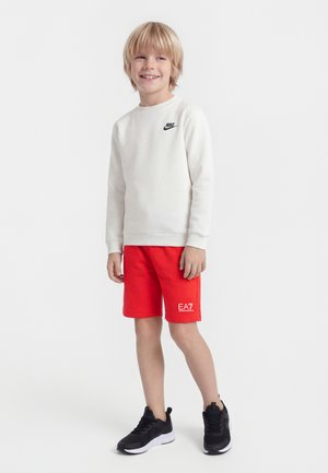 Usmievajúci sa mladý chlapec s blond vlasmi stojí oblečený v bielom mikinovom pulóvri Nike, červených šortkách EA7 a čiernych teniskách na jednoduchom pozadí.