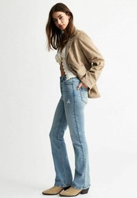 Jeans acampanados de cintura alta en azul claro con un detalle deshilachado. Combinados con un top de punto color crema y una blazer de ante color marrón. Botines de tacón color marrón.