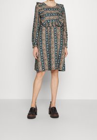 Šaty s květinovým vzorem v tmavě zelené barvě, s volánovým výstřihem a dlouhými rukávy, s plisováním a uvolněným střihem. Černé robustní mokasíny dobře ladí.