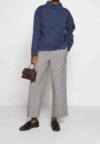 Pullover navy con colletto alto, abbinato a pantaloni larghi grigio a quadri. Borsa nera con finitura testurizzata e accessori in oro. Scarpe nere.