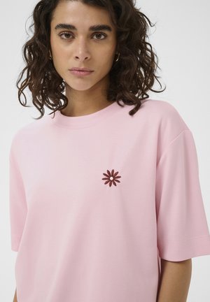 Mujer con cabello rizado y oscuro que lleva una camiseta rosa claro con una pequeña flor de color rojo oscuro bordada en el pecho izquierdo.