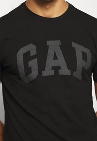 Camiseta de algodón negra con un gran logo gráfico en gris "GAP" en una fuente bold y de bloque. Escote redondo; mangas cortas. Textura suave.