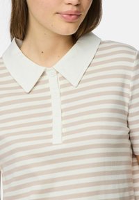 Svītrainā polo krekls gaiši bēšā un baltā krāsā, ar plašu baltu apkakli, īsām piedurknēm un piecām baltām pogām pie pogas. 