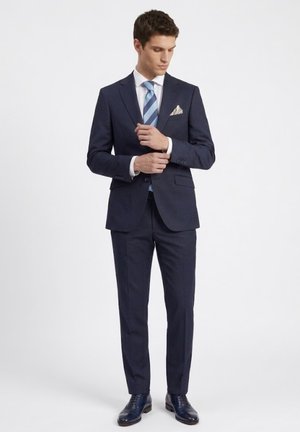 Hombre ajustándose los gemelos, vestido con un traje azul marino, camisa blanca, corbata azul con rayas, pañuelo de bolsillo y zapatos de vestir azul marino pulidos.