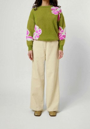 Kobieta ubrana w oliwkowozielony sweter z różowo-białymi kwiatowymi wzorami, kremowe spodnie o szerokich nogawkach i brązowe buty na kwadratowym nosku, stojąca.