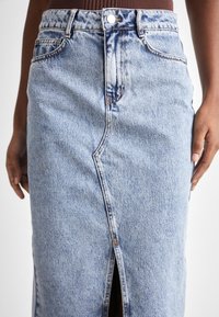 Denimová sukně ve světle modré barvě s výřezem v přední části. Mezi její rysy patří vysoký pas, zapínání na knoflík a vybledlá textura se švy.