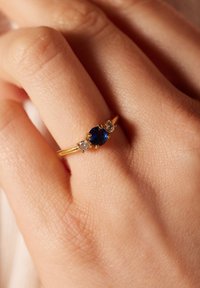 Gouden ring met een centrale ronde blauwe saffier, geflankeerd door twee kleinere heldere stenen, geplaatst tegen een getextureerde achtergronde.