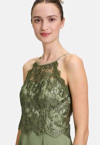 Oliwkowa sukienka z przezroczystym, haftowanym gorsetem, zdobionym kwiatowymi wzorami i cekinami. Ramiączka spaghetti podkreślają dekolt.