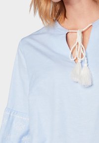 Helle blaue Bluse mit einem Schlüsselloch-Ausschnitt, ausgestattet mit weißen Quasten und gestickten Akzenten an den Ärmeln. Weicher Stoff und lockere Passform.
