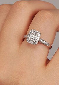 Quadratisch gefasster diamantener Ring mit einem smaragdgeschliffenen Centerstein, umgeben von kleineren Diamanten, in einem polierten Silberband.