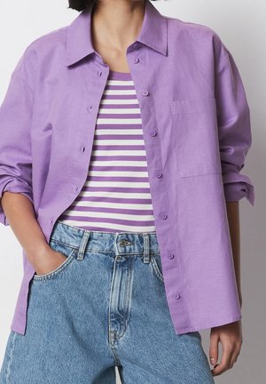 Personne portant une chemise lavande à boutons sur un débardeur rayé violet et blanc, associée à un jean bleu taille haute.