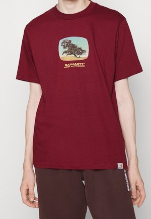 Osoba ubrana w bordowy t-shirt Carhartt z nadrukiem drzewa i napisem „Carhartt Work in Progress” na piersi, w połączeniu z ciemnobrązowymi spodniami.