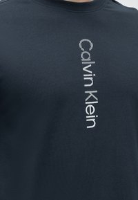 Σκούρο μπλε μπλούζα με λευκό κάθετο λογότυπο "Calvin Klein" στην αριστερή πλευρά του στήθους, φτιαγμένο από διακεκομμένες γραμμές.