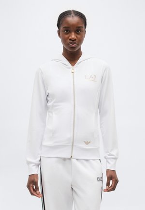 Naine, kes kannab valget lukkuga kapuutsiga pusa ja sobivaid valgeid pükse musta ja rohelise triibuga, seisab ühetoonilise tausta ees.