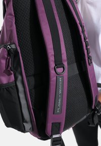 Zaino viola con dettagli neri, dotato di spallacci imbottiti in rete, tasche con cerniera e un logo "PLANET WATER" su un pannello testurizzato.