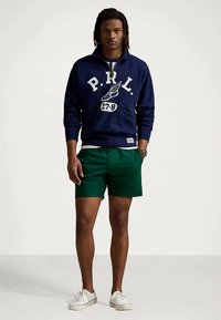 Sweatshirt azul-marinho com logo branco e bolso canguru, combinado com calções verdes. Calçado é ténis brancos. Fundo neutro.