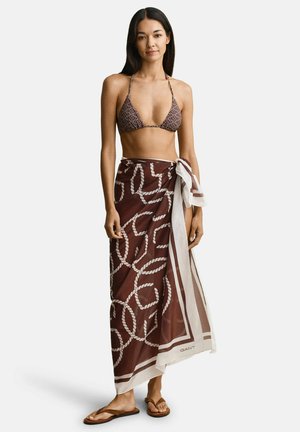 Frau trägt ein braun gemustertes Bikini-Oberteil und einen passenden braunen Sarong, der an der Taille gebunden ist, steht barfuß vor weißem Hintergrund.