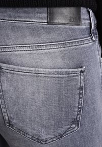 Jeans de denim gris con una textura suave, que presentan un bolsillo trasero y una etiqueta de cuero negro grabada con "RIVER ISLAND" en la cinturilla.